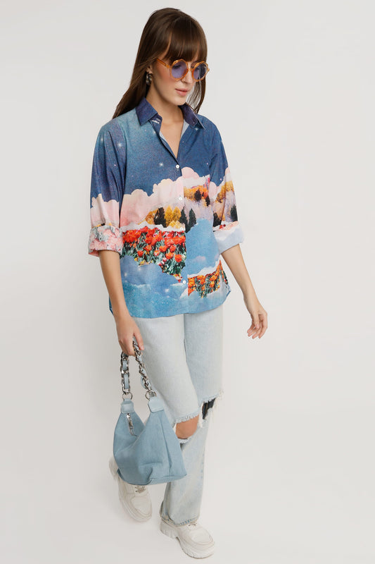A woman modeling a blue short-sleeve button-up shirt with a colorful landscape print, paired with light-wash ripped jeans, white sneakers, and a light blue handbag, against a plain white studio background.