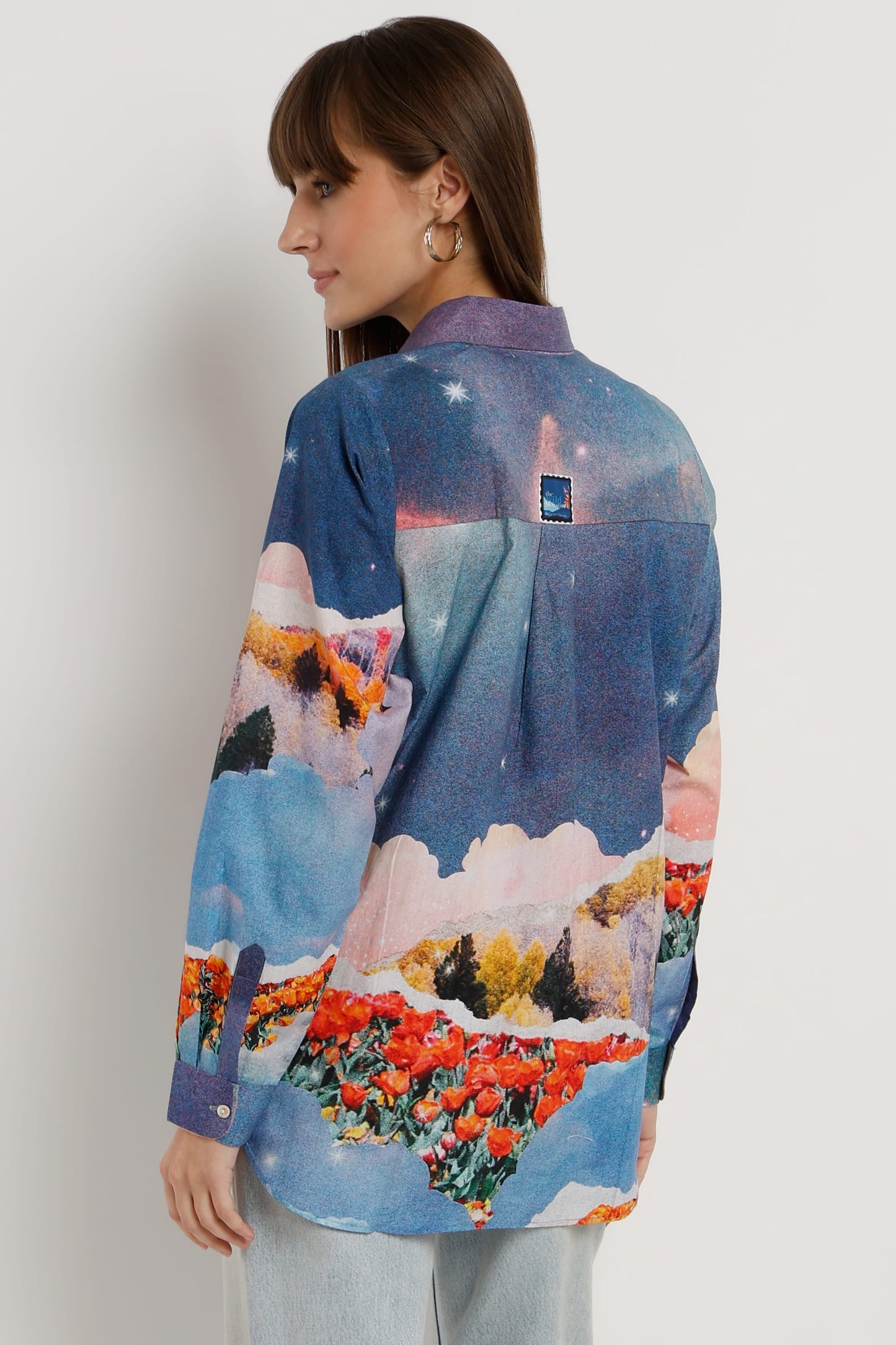 A woman shown from the back wearing a long-sleeve button-up shirt with a colorful landscape print featuring blue skies, mountains, and red flowers, paired with light gray jeans against a plain light gray background.