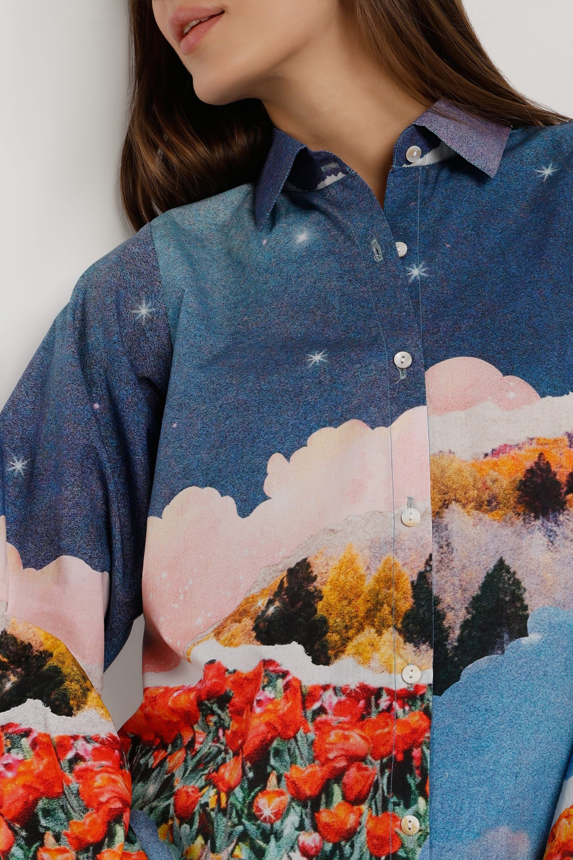A close-up of a woman wearing a button-up shirt with a colorful landscape print featuring blue sky, pink clouds, orange trees, and red flowers.