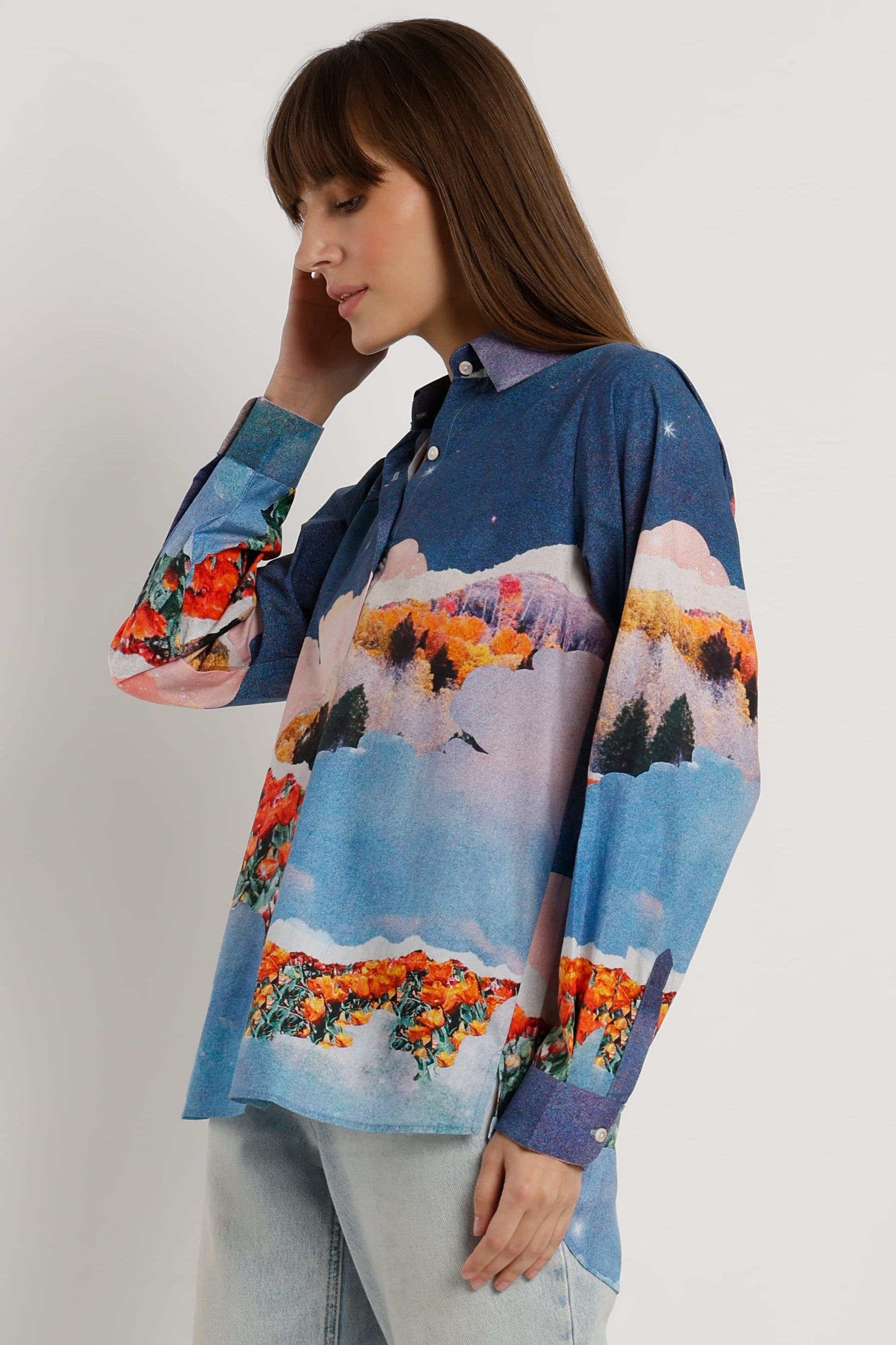 A woman wearing a long-sleeve button-up shirt with a colorful landscape print, paired with light-wash jeans, photographed against a plain light gray studio background.