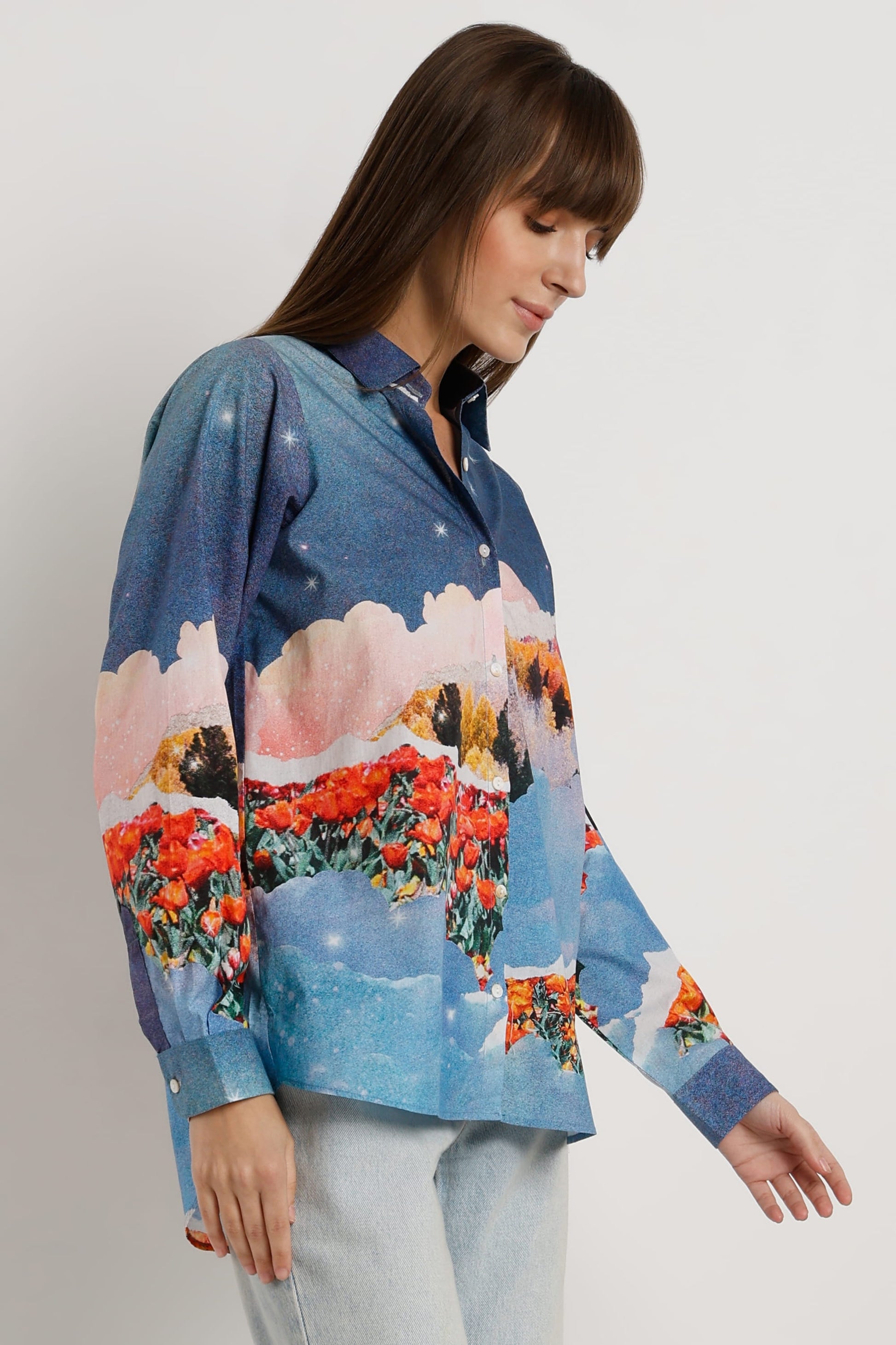 A woman wearing a long-sleeve button-up shirt with a colorful landscape print featuring blue skies, pink clouds, and red flowers, paired with light blue jeans against a plain light gray background.