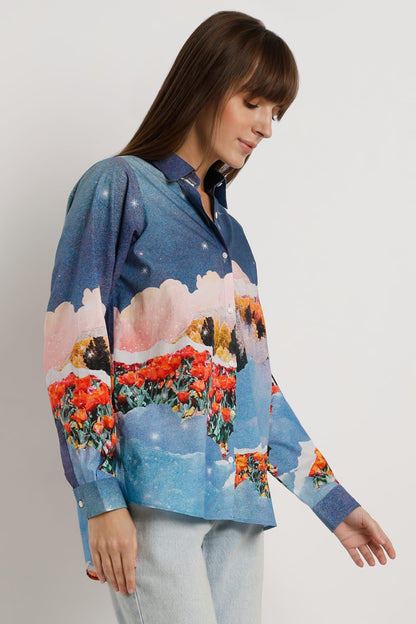 A woman wearing a long-sleeve button-up shirt with a colorful landscape print featuring blue skies, pink clouds, and red flowers, paired with light blue jeans against a plain light gray background.