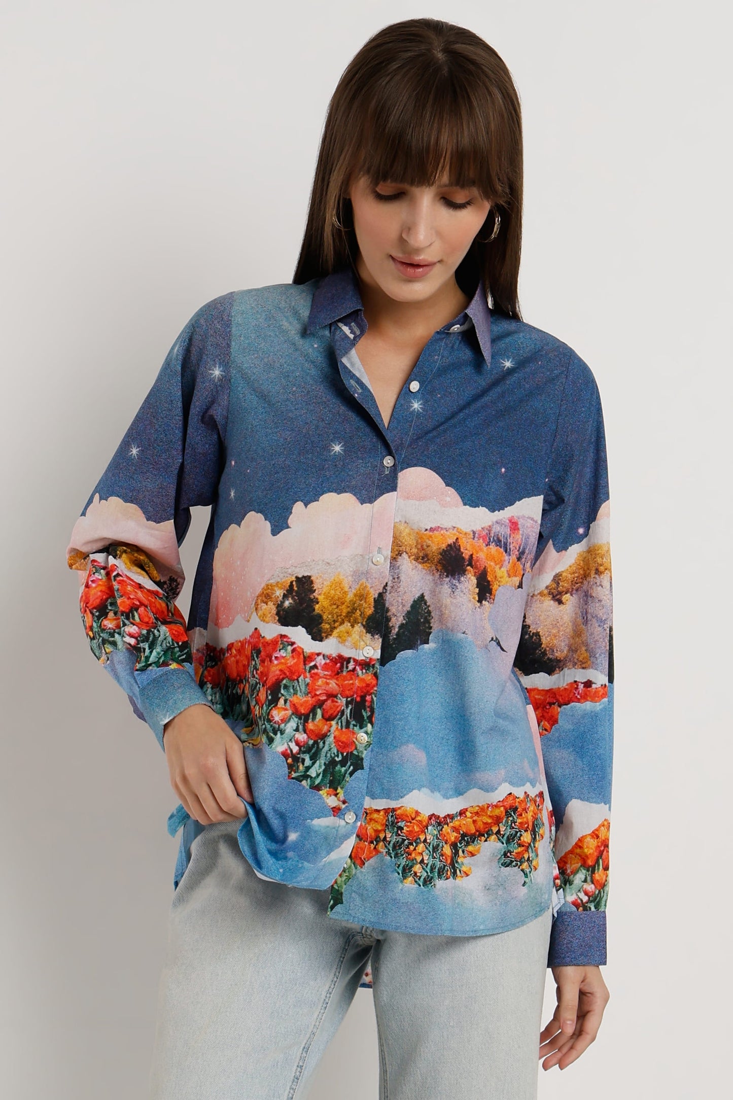 A woman wearing a long-sleeve button-up shirt with a colorful landscape print featuring blue sky, clouds, and red/orange flowers, paired with light gray jeans, against a plain light gray background.