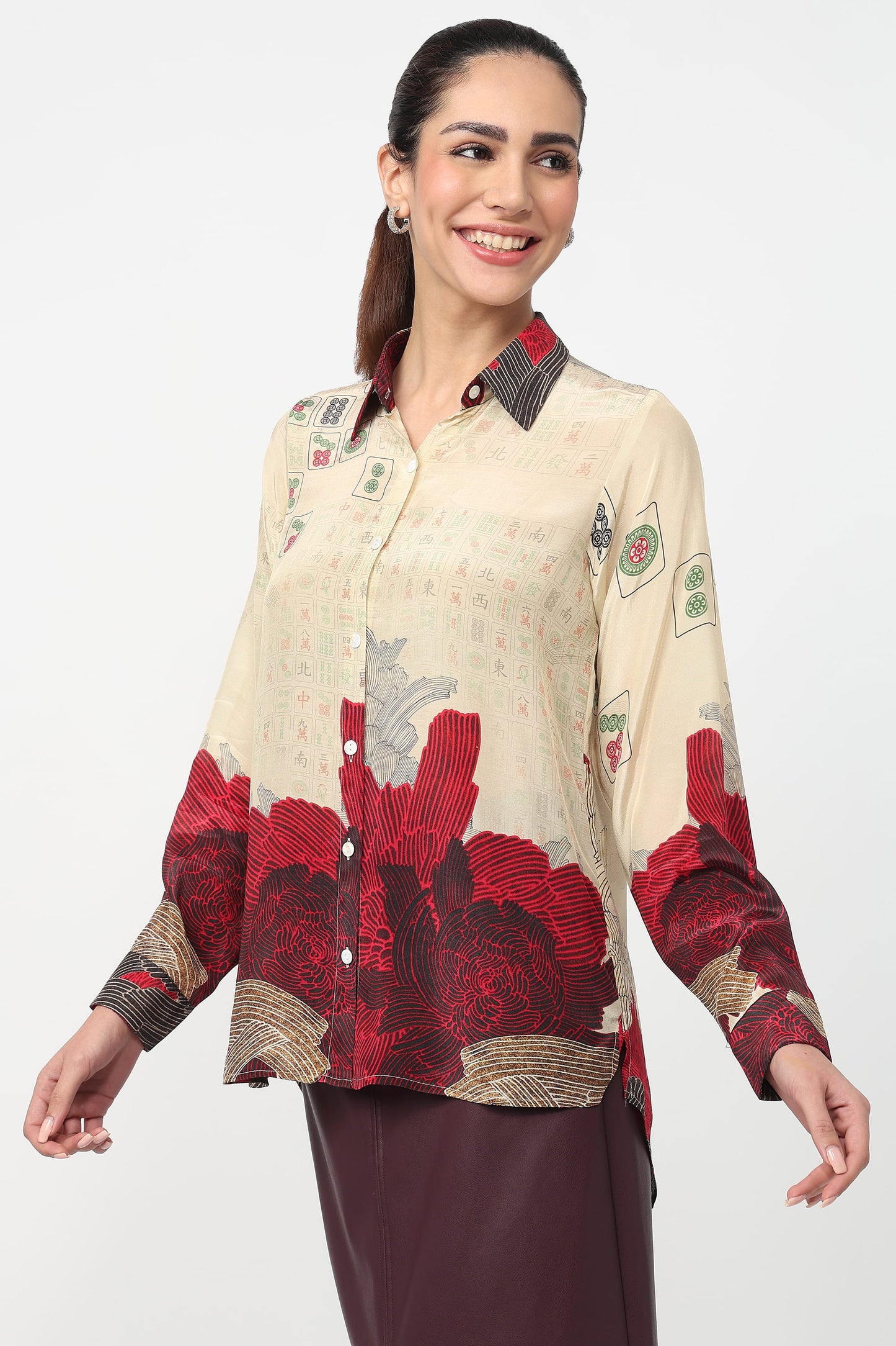 A woman modeling a long-sleeve button-up blouse with a beige base and a bold red floral print concentrated on the lower half, paired with dark brown pants against a light gray studio background.
