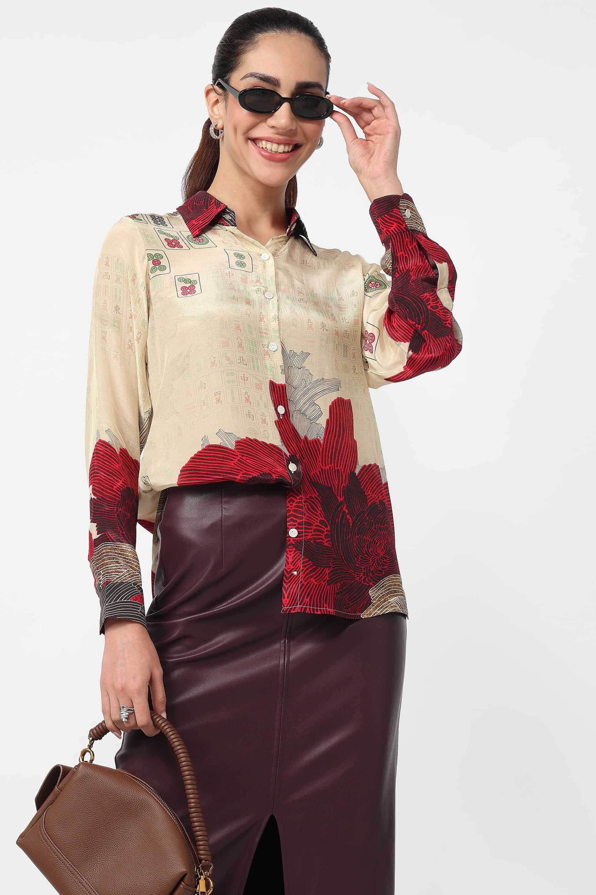 Woman wearing a patterned shirt with red and beige design, holding sunglasses, and standing against a white background.