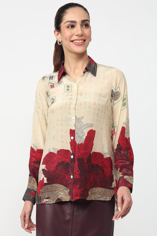 A woman modeling a long-sleeve button-up shirt with a beige base, red floral print, and small graphic details, paired with a dark brown skirt against a light gray studio background.