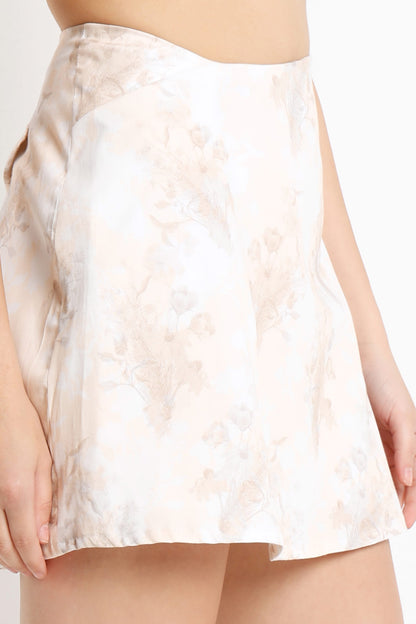 A close-up of a model wearing a short, flared satin skirt with a soft beige floral jacquard pattern on a white base.