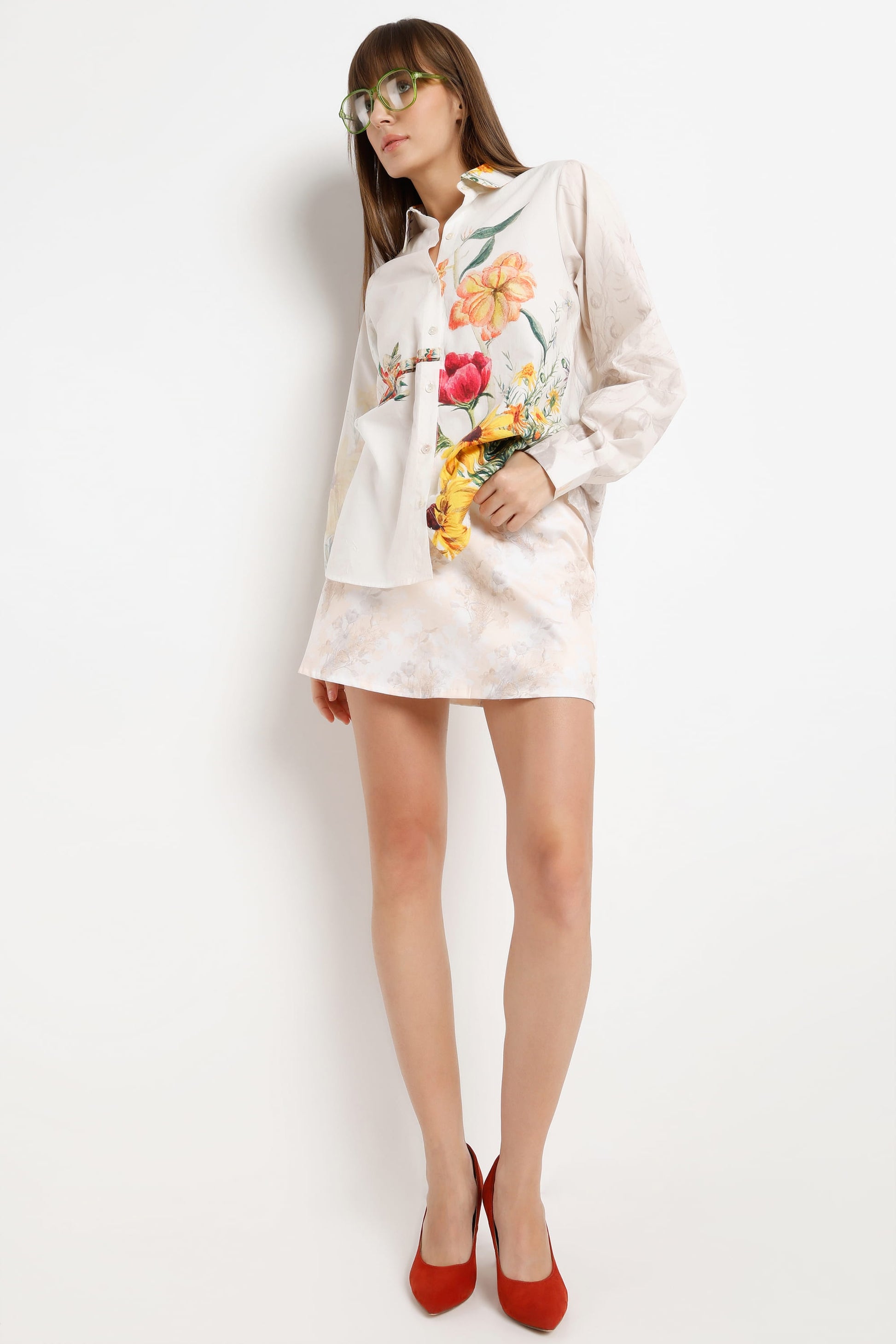 A woman modeling a white satin shirt jacket with colorful floral embroidery, paired with red high heels and green-tinted sunglasses, against a plain white background.