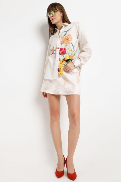 A woman modeling a white satin shirt jacket with colorful floral embroidery, paired with red high heels and green-tinted sunglasses, against a plain white background.