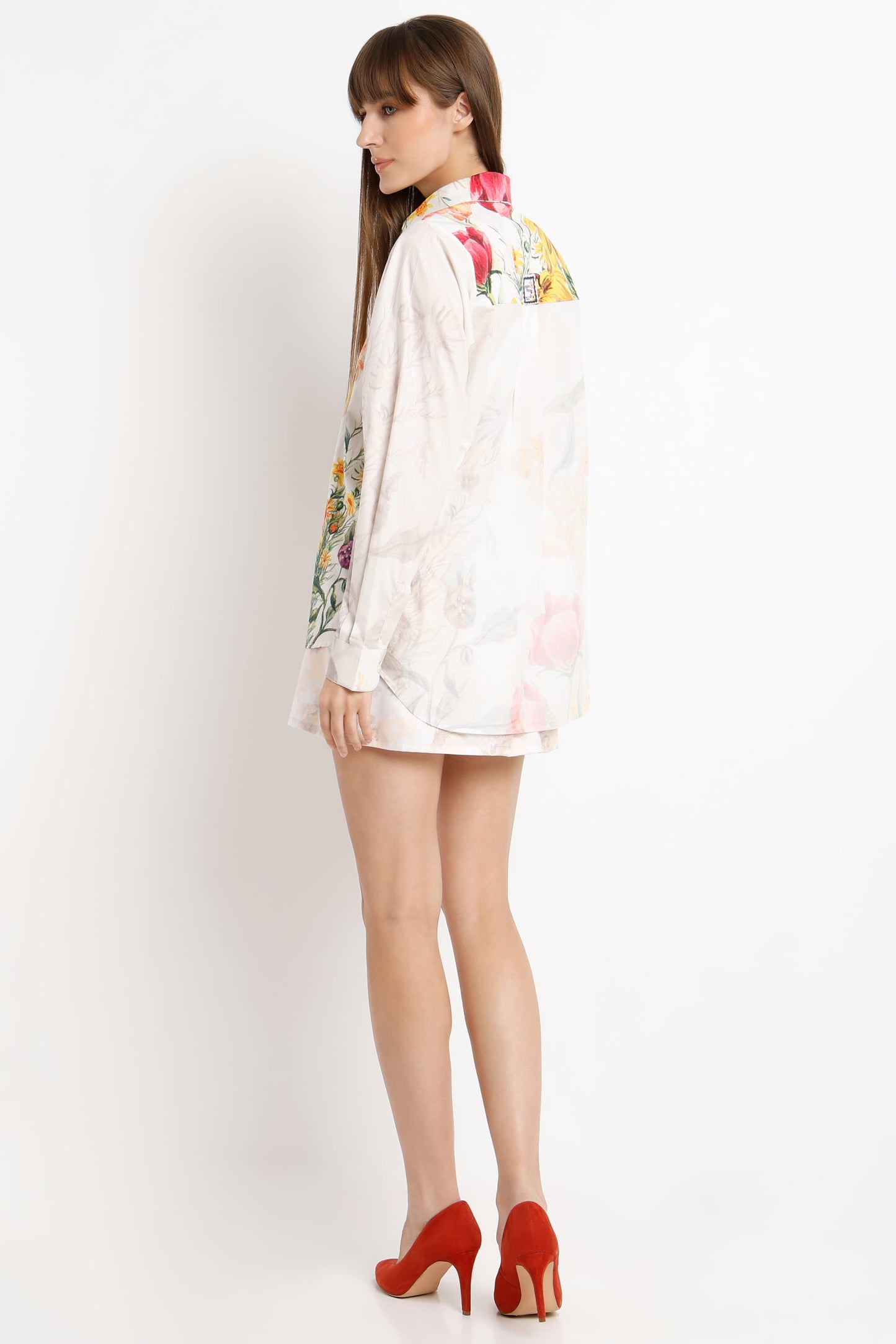 A woman shown from the back wearing a white long-sleeve shirt dress with colorful floral embroidery on the collar and sleeves, paired with red high heels, against a plain white background.