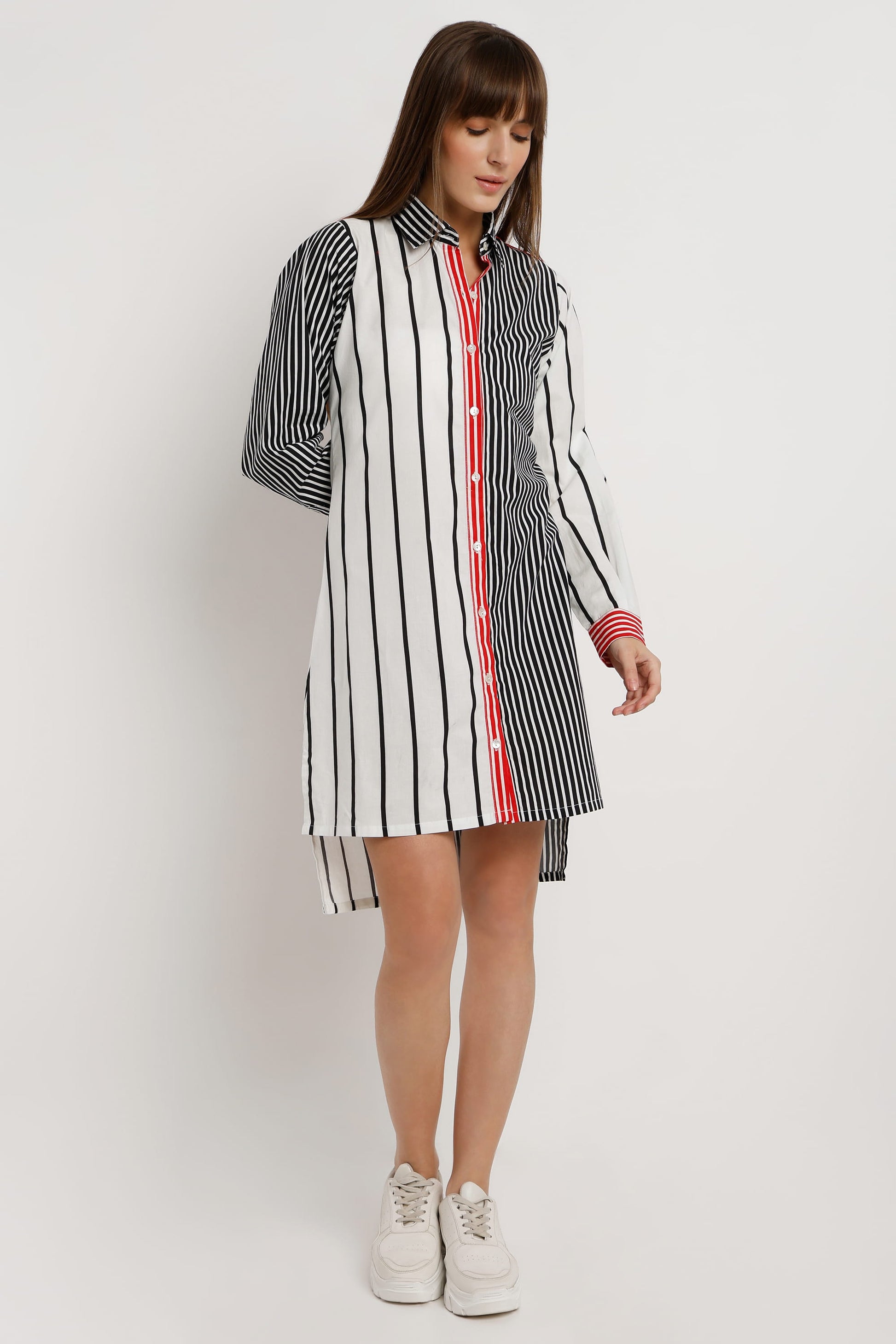 A woman wearing a long-sleeve shirt dress with vertical stripes in black and white, featuring a solid red front placket and red striped cuffs, paired with white sneakers against a light white studio background.