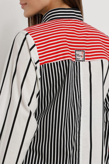A woman wearing a long-sleeve shirt dress with vertical stripes in black and white, featuring a solid red front placket and red striped cuffs, paired with white sneakers against a light white studio background.