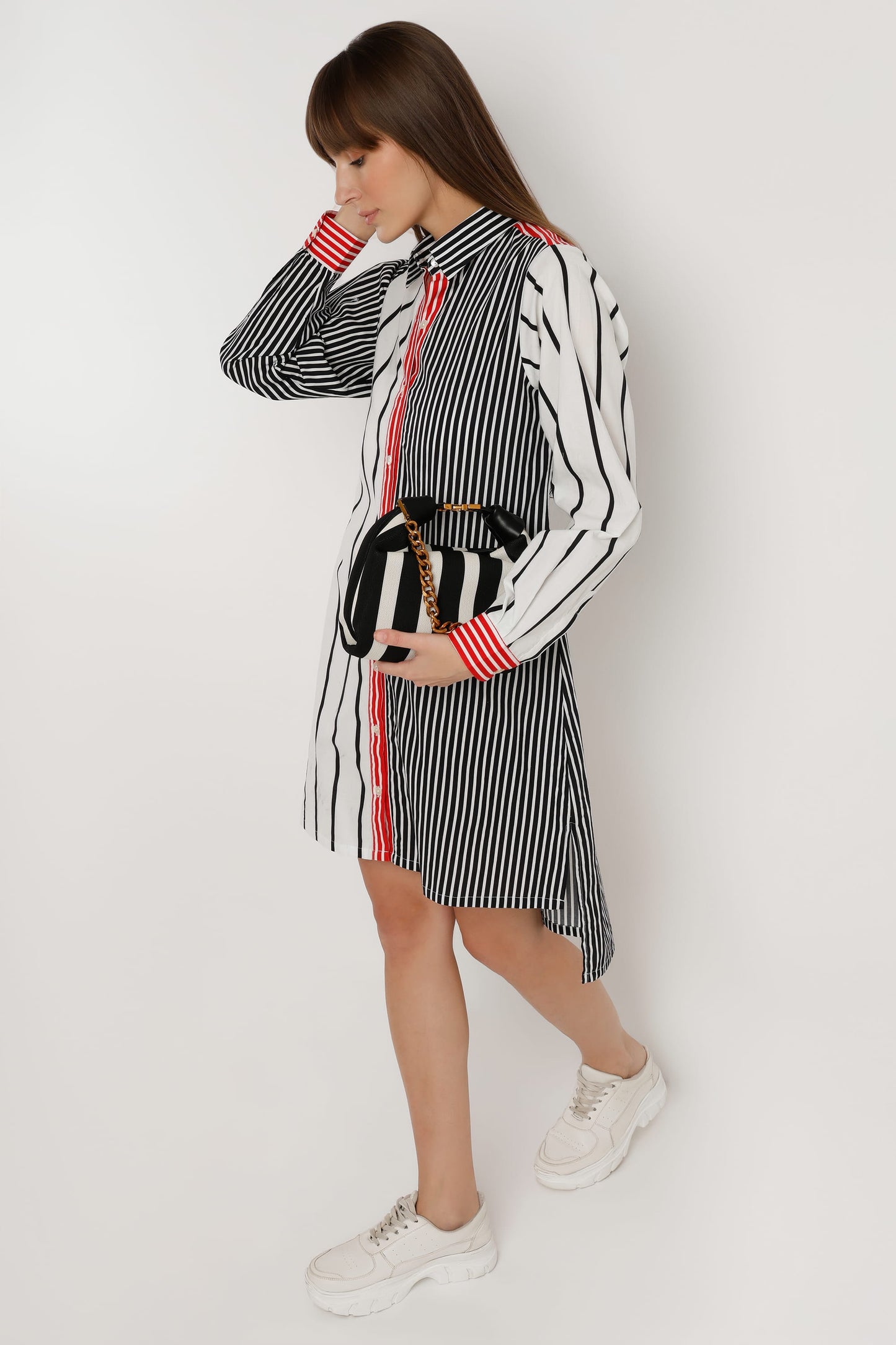 A woman wearing a long-sleeve shirt dress with vertical stripes in black and white, featuring a solid red front placket and red striped cuffs, paired with white sneakers against a light white studio background.