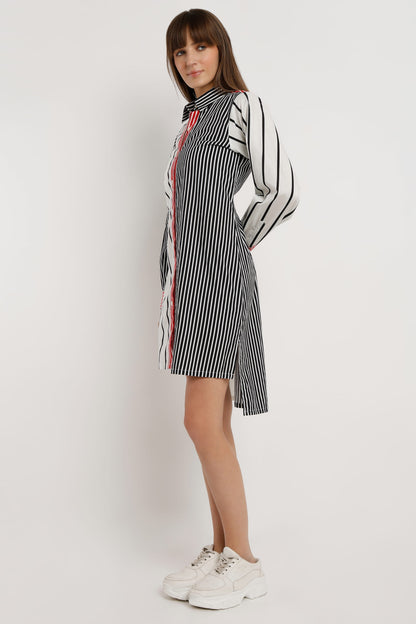 A woman wearing a long-sleeve shirt dress with vertical stripes in black and white, featuring a solid red front placket and red striped cuffs, paired with white sneakers against a light white studio background.
