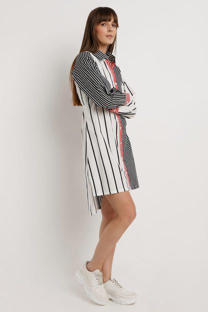 A woman wearing a long-sleeve shirt dress with vertical stripes in black and white, featuring a solid red front placket and red striped cuffs, paired with white sneakers against a light white studio background.