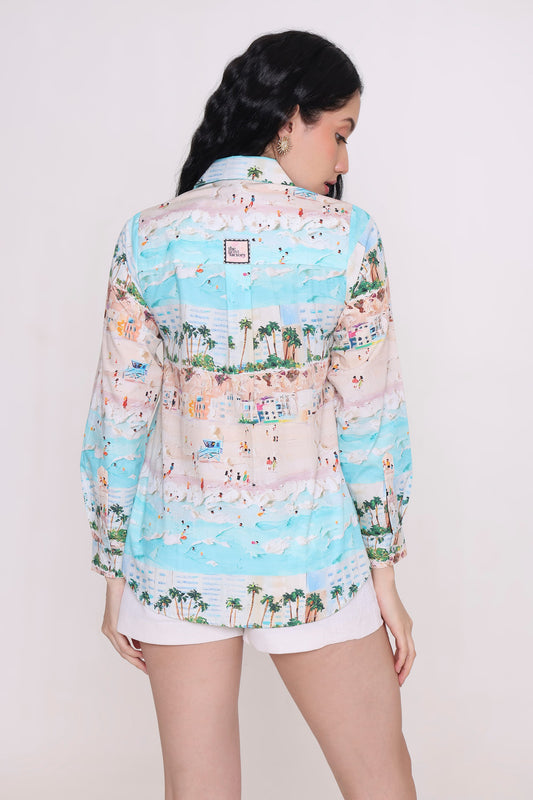 A woman shown from the back wearing a long-sleeve button-up shirt with a colorful beach scene print, paired with white shorts, against a plain light background.