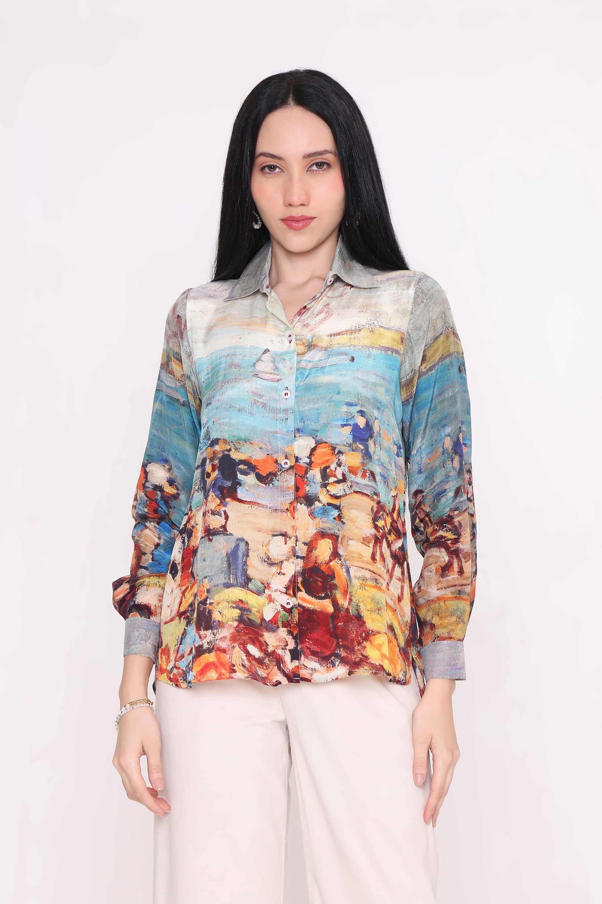 A woman modeling a long-sleeve button-up shirt with a colorful, painterly beach scene print, paired with light beige trousers, against a plain white background.