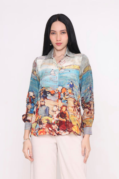 A woman modeling a long-sleeve button-up shirt with a colorful, painterly beach scene print, paired with light beige trousers, against a plain white background.