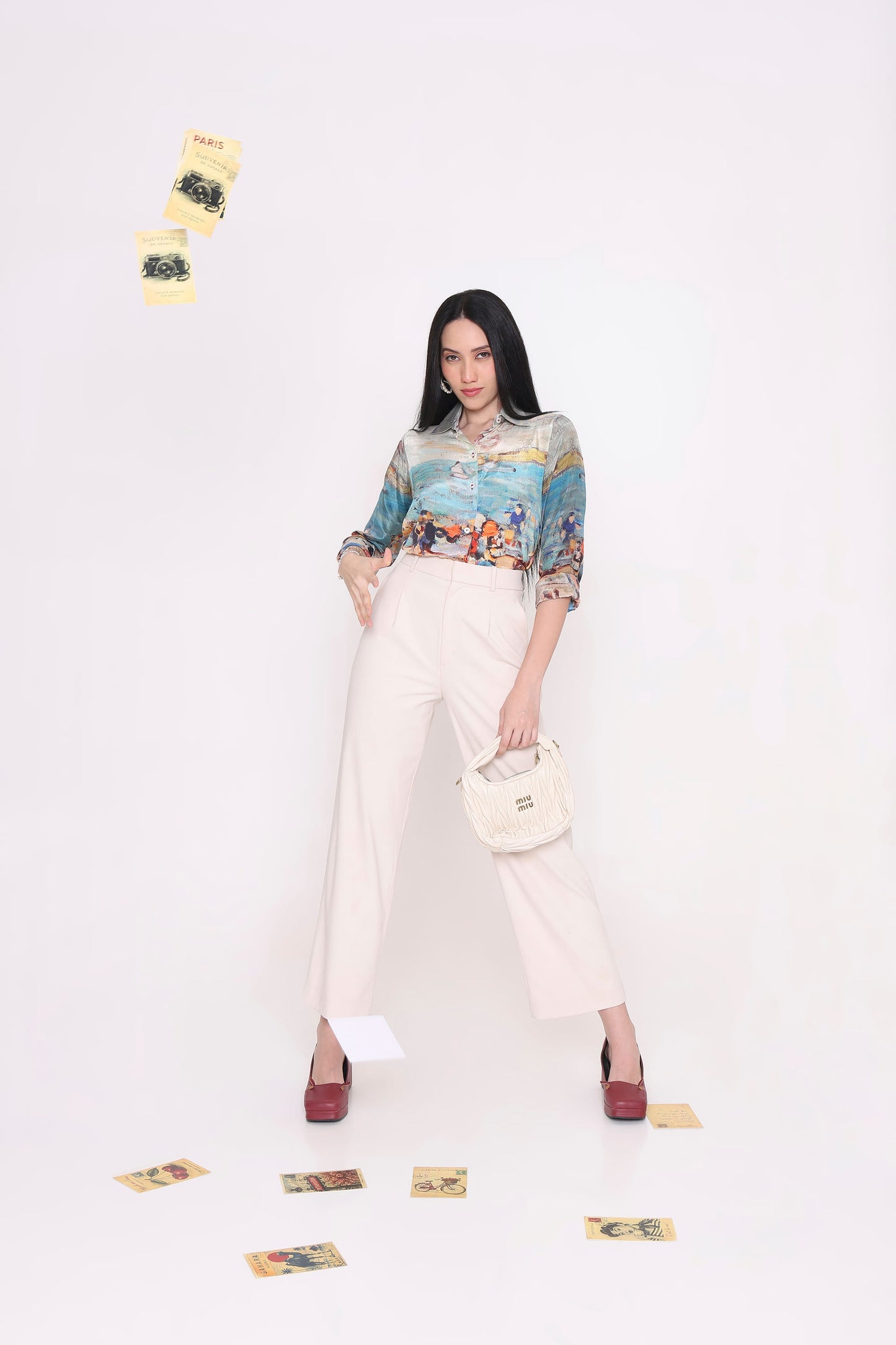A woman stands against a plain white studio background wearing a colorful patterned button-up blouse, cream wide-leg trousers, and burgundy heels, while holding a small cream handbag. Several small cards are scattered on the floor and floating in the air.
