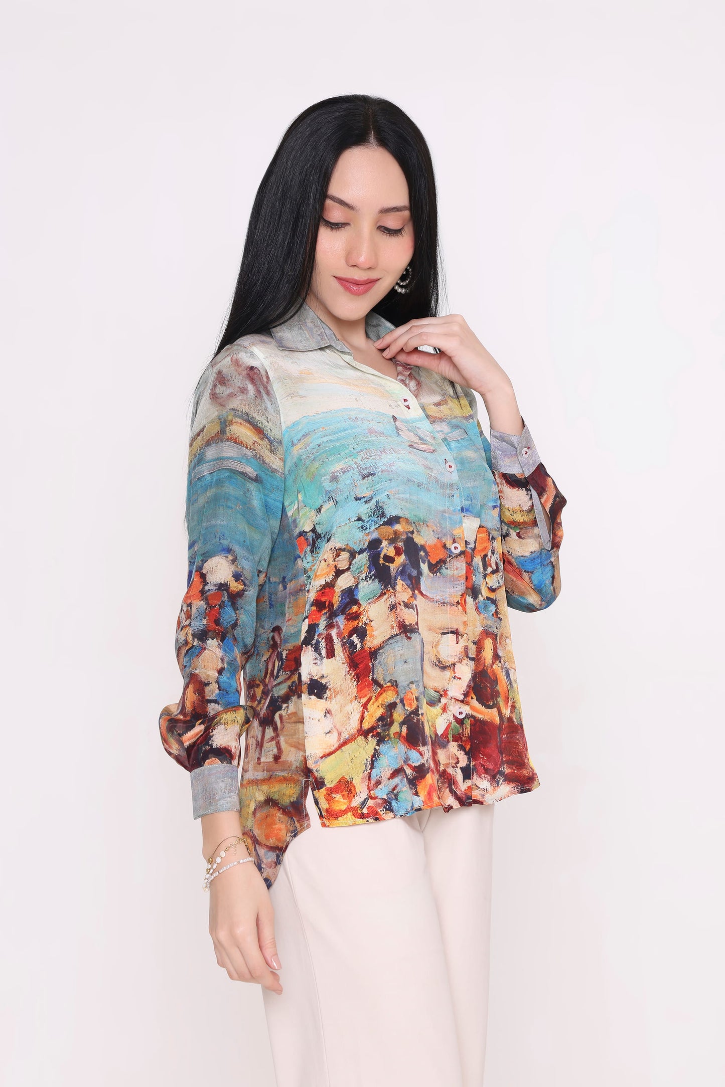 A woman modeling a long-sleeve button-up blouse with a colorful, painterly abstract print, paired with light beige trousers, against a plain white studio background.