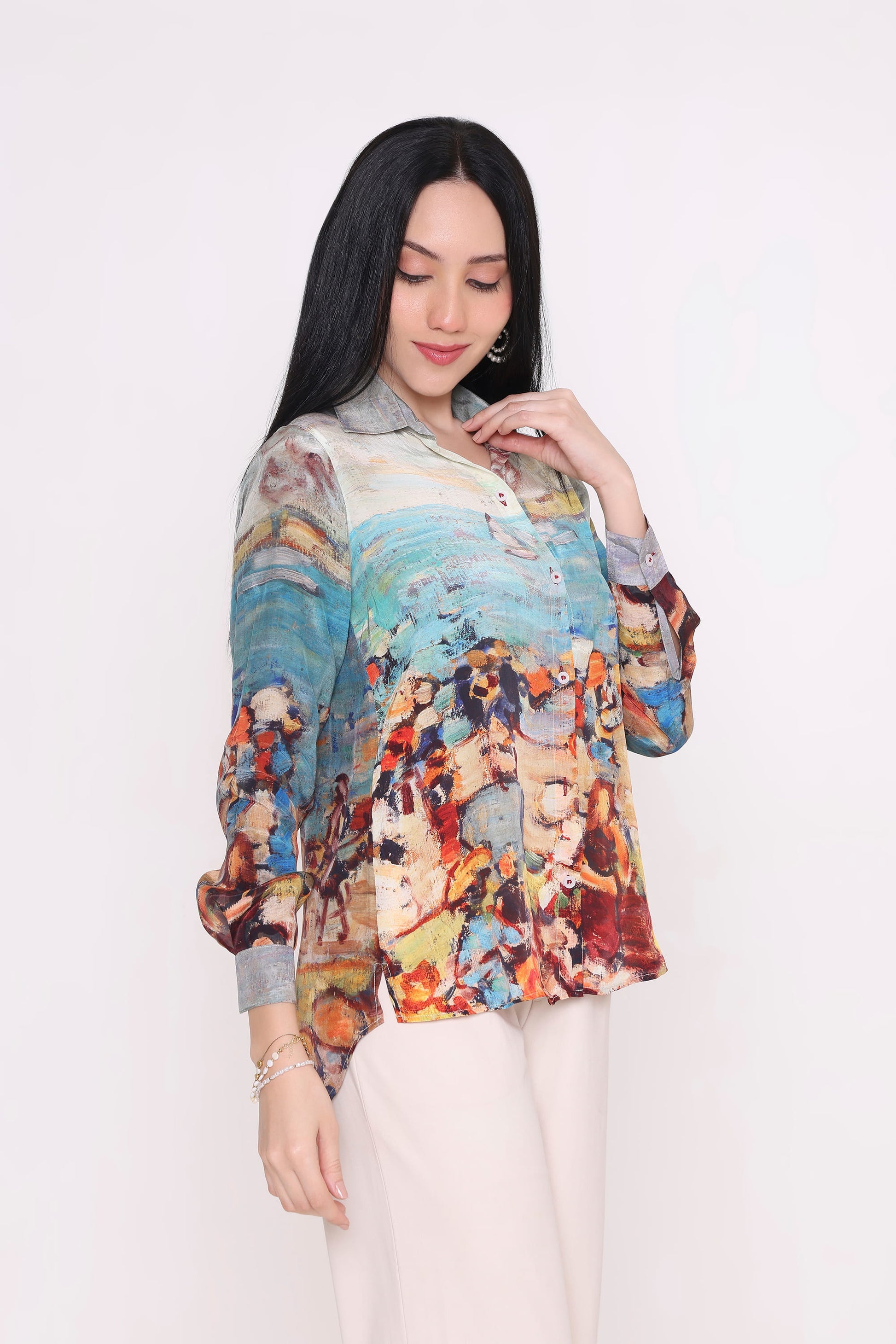 A woman modeling a long-sleeve button-up blouse with a colorful, painterly abstract print, paired with light beige trousers, against a plain white studio background.