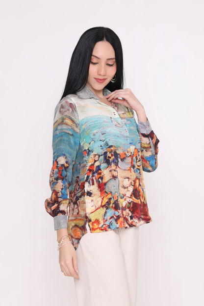 A woman modeling a long-sleeve button-up blouse with a colorful, painterly abstract print, paired with light beige trousers, against a plain white studio background.