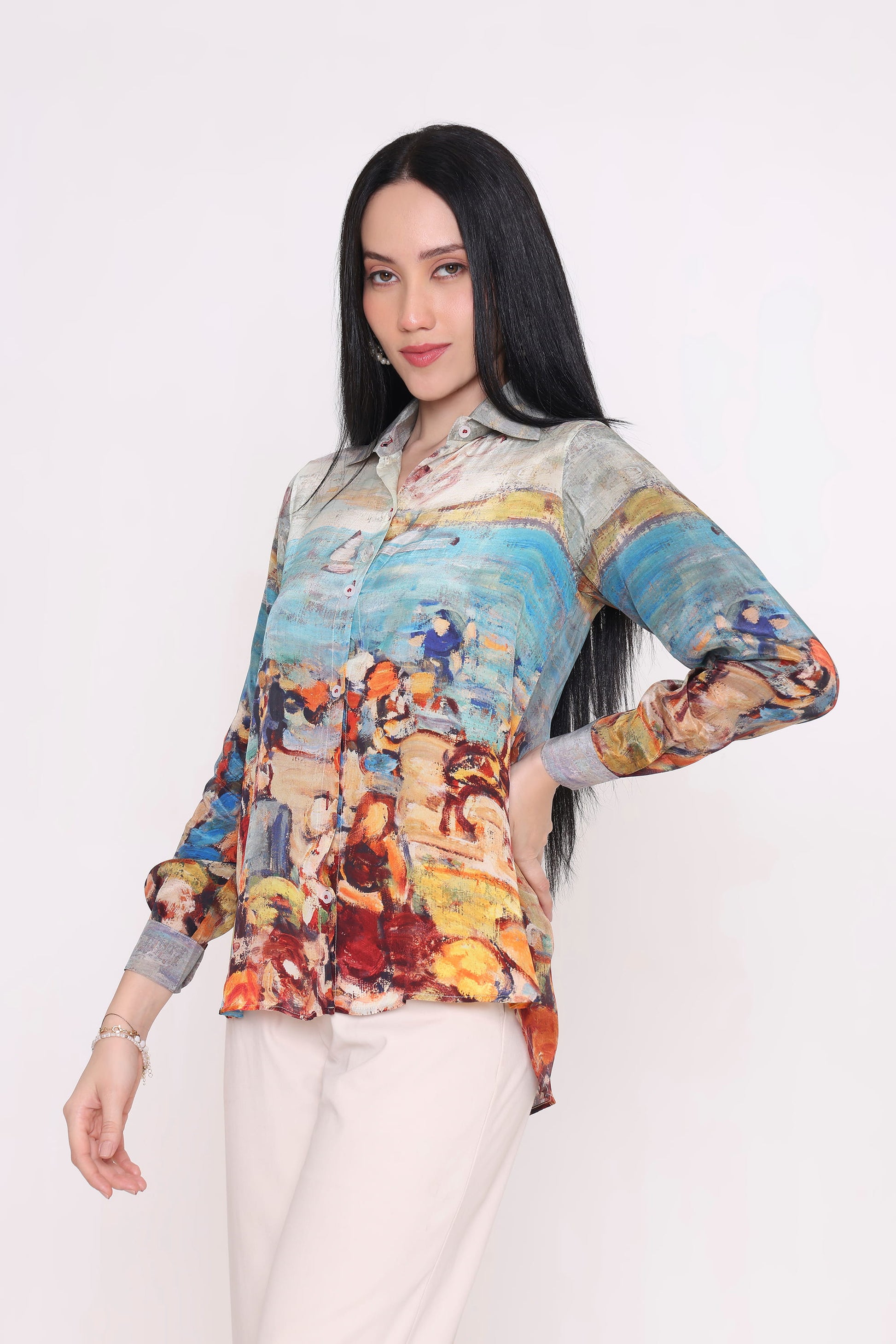 A woman modeling a long-sleeve button-up shirt with a colorful, painterly beach scene print, paired with light beige trousers, against a plain white studio background.
