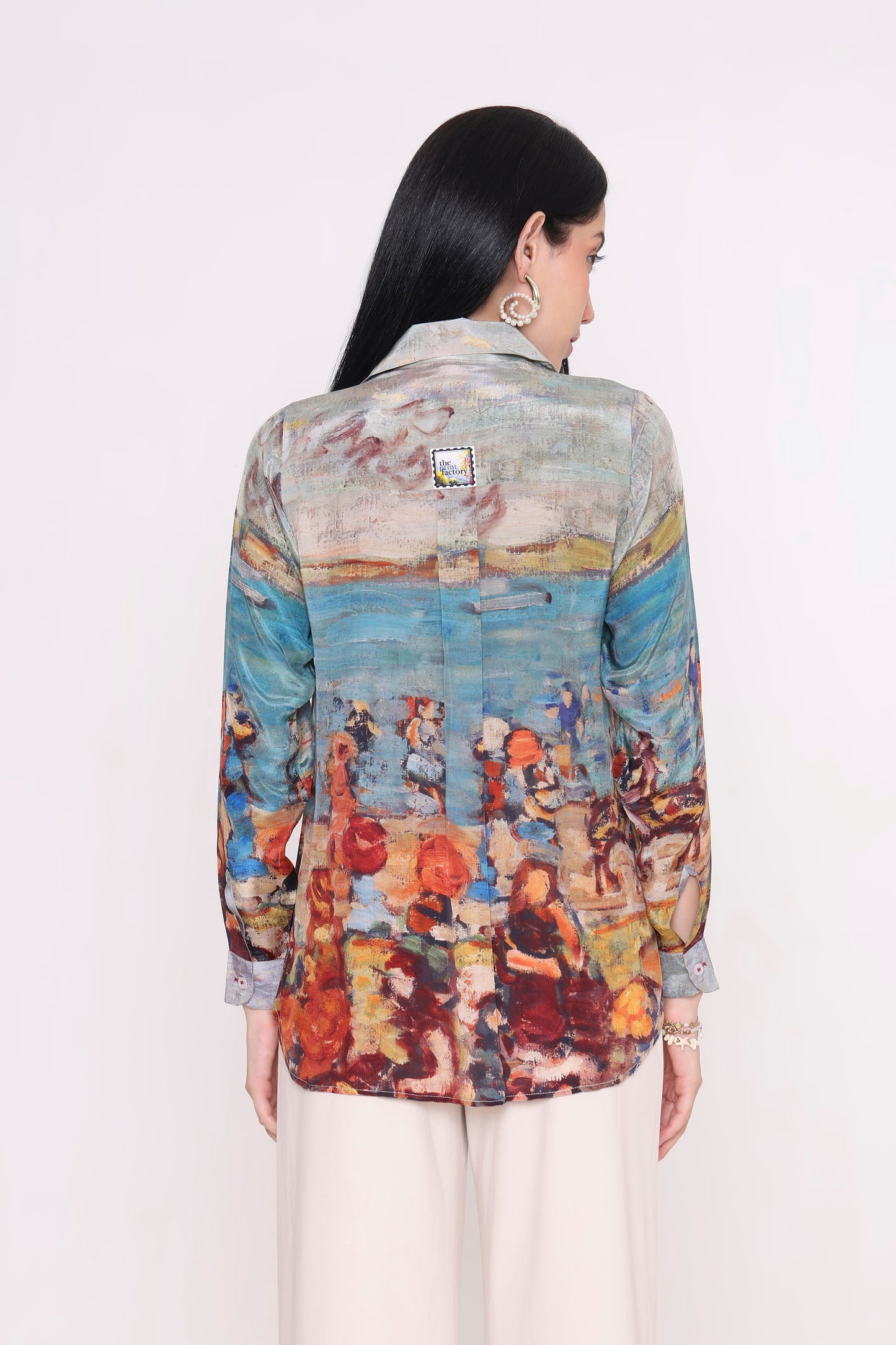A woman shown from the back wearing a long-sleeve button-up shirt with a colorful, painterly beach scene print, paired with light beige trousers, against a plain white background.