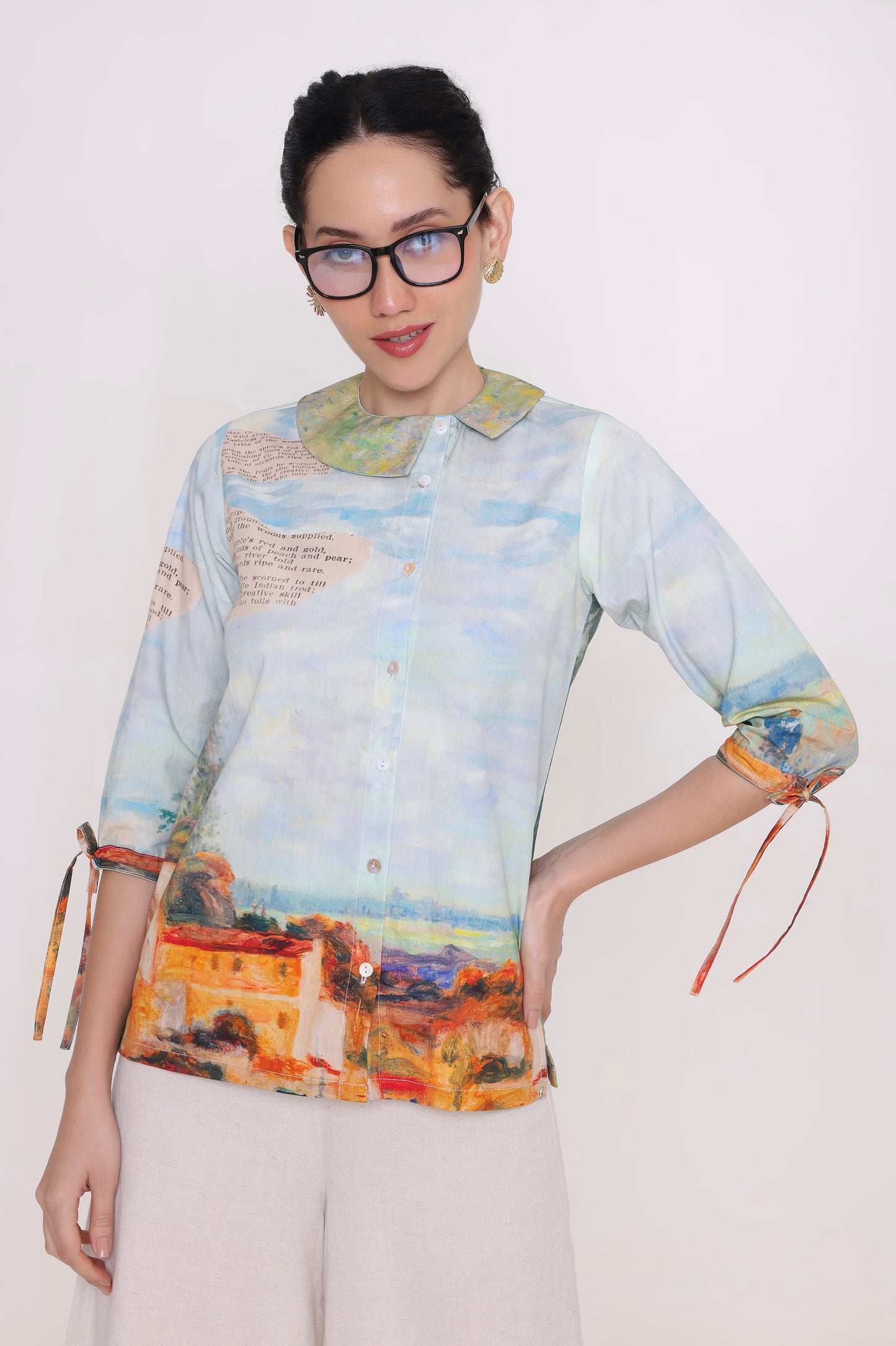A woman wearing a light blue and white printed button-up shirt with a scenic landscape design, a green collar, and rolled sleeves with tie details, paired with white pants against a plain white background.