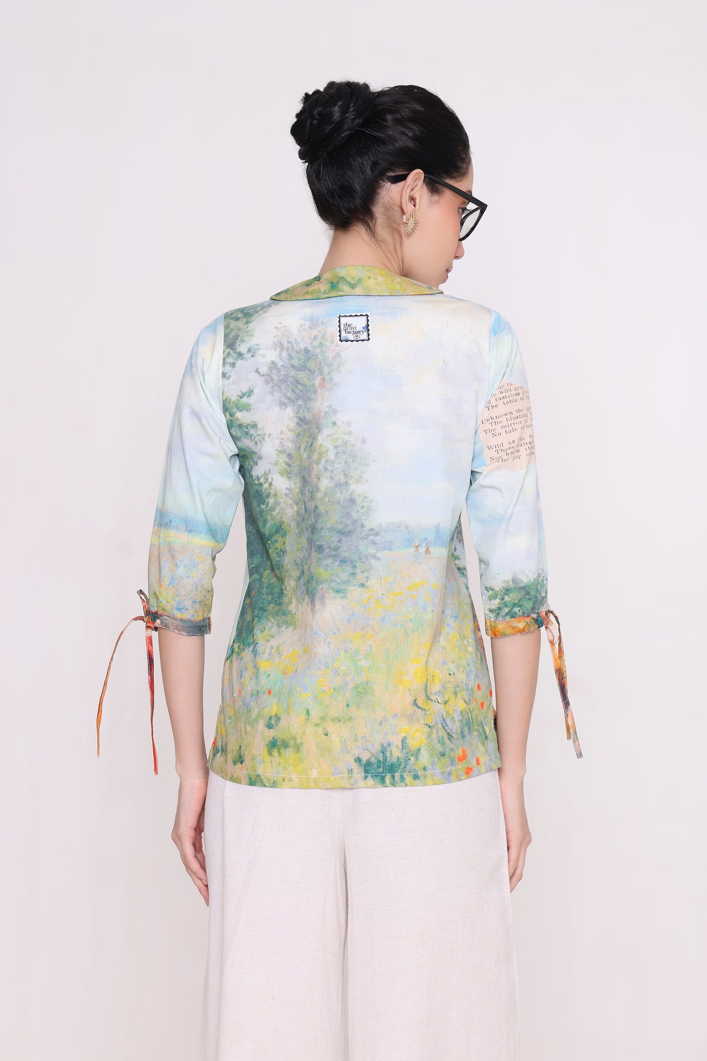 A woman viewed from the back wearing a light blue and yellow painterly landscape print shirt with three-quarter sleeves and side ties, paired with off-white pants, against a plain light background.