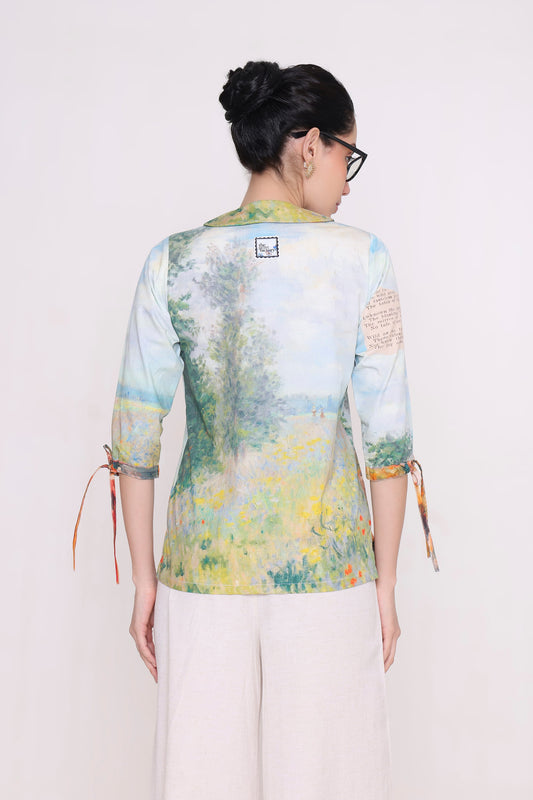 A woman viewed from the back wearing a light blue and yellow painterly landscape print shirt with three-quarter sleeves and side ties, paired with off-white pants, against a plain light background.