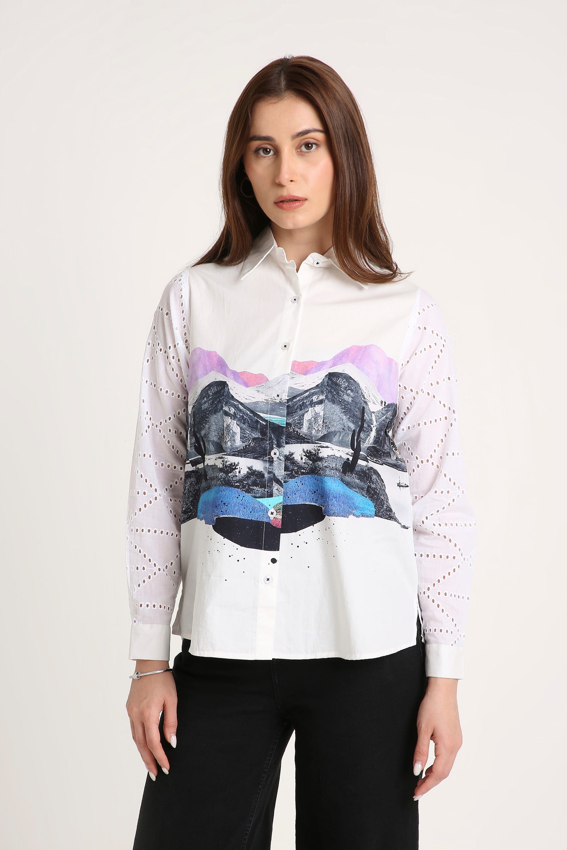 A woman wearing a white long-sleeve button-up shirt with a colourful abstract landscape graphic across the front and sheer white dotted mesh sleeves, paired with black pants against a light grey studio background.