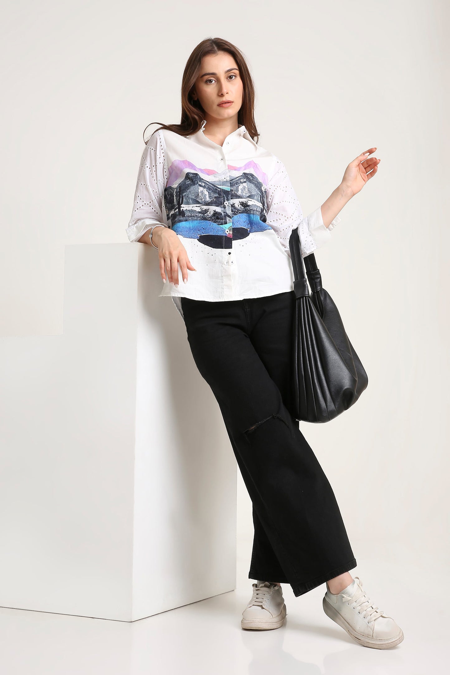 A woman wearing a white long-sleeve button-up shirt with a colourful graphic print on the front, paired with black wide-leg pants, white sneakers, and a black shoulder bag, standing next to a white display block.