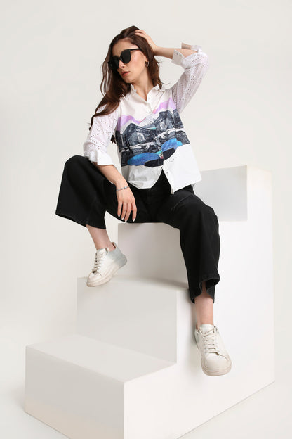 Woman sitting on a white block wearing a white shirt with a colorful graphic, black pants, and white sneakers.