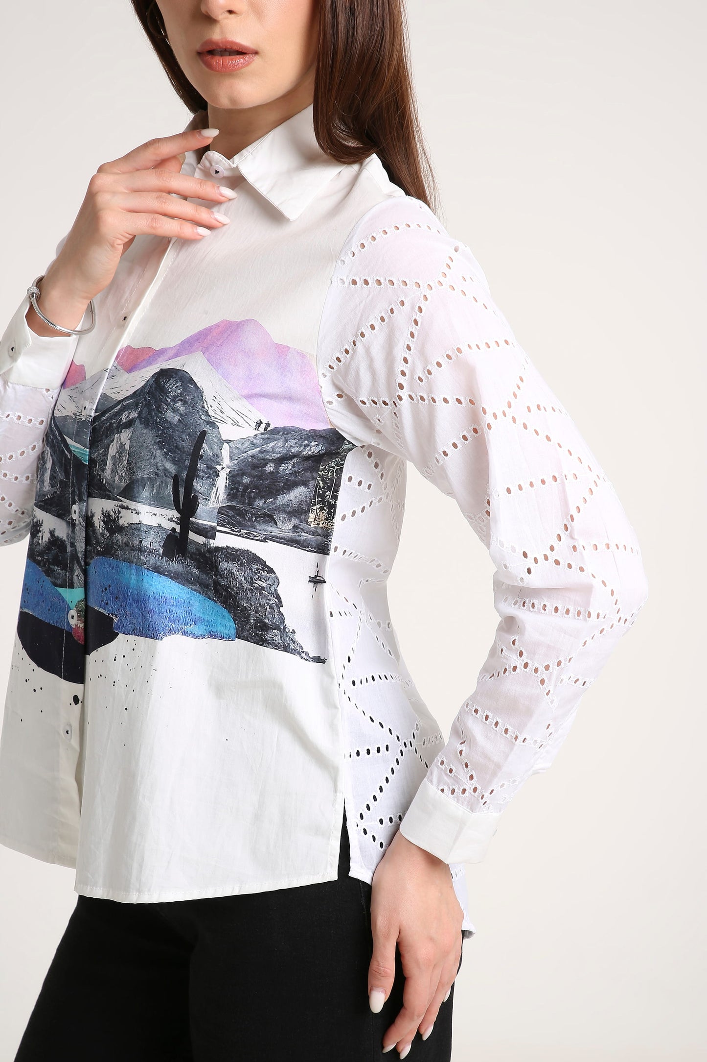 A woman wearing a white long-sleeve button-up shirt with a colorful mountain landscape print on the front and sheer geometric eyelet sleeves, paired with black pants against a light beige studio background.