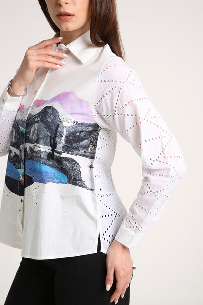 A woman wearing a white long-sleeve button-up shirt with a colorful mountain landscape print on the front and sheer geometric eyelet sleeves, paired with black pants against a light beige studio background.
