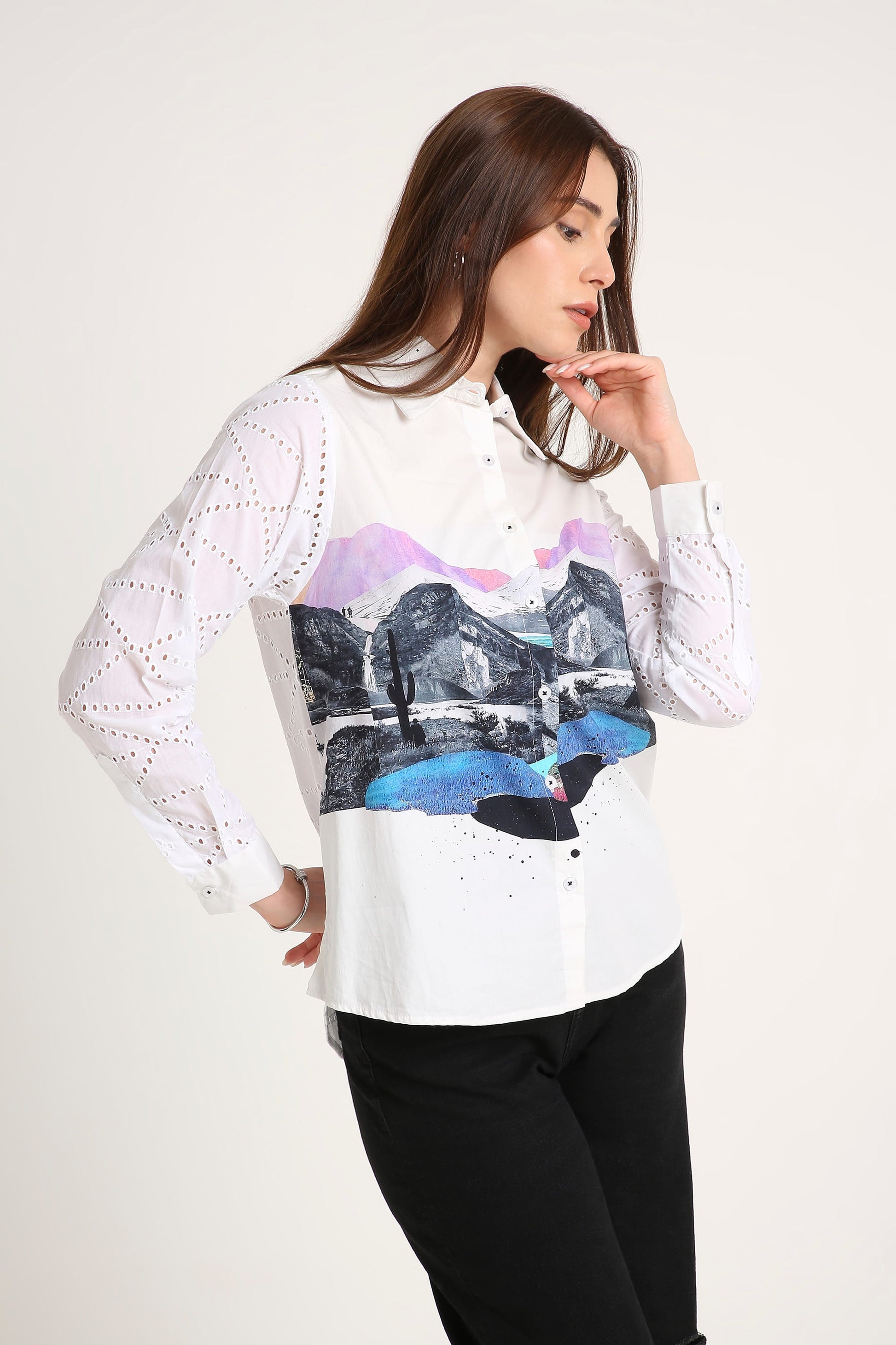A woman wearing a white long-sleeve button-up shirt with eyelet lace sleeves and a colourful front graphic print of a mountain landscape, paired with black pants against a plain light background.