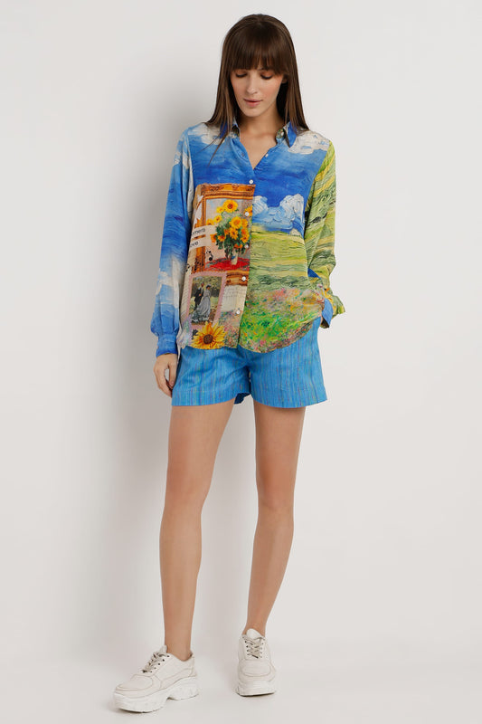 A woman modeling a long-sleeve button-up shirt and matching shorts set. The set features a vibrant, multicolor landscape art print on the shirt and a blue textured stripe pattern on the shorts, paired with white sneakers against a plain light gray studio background.