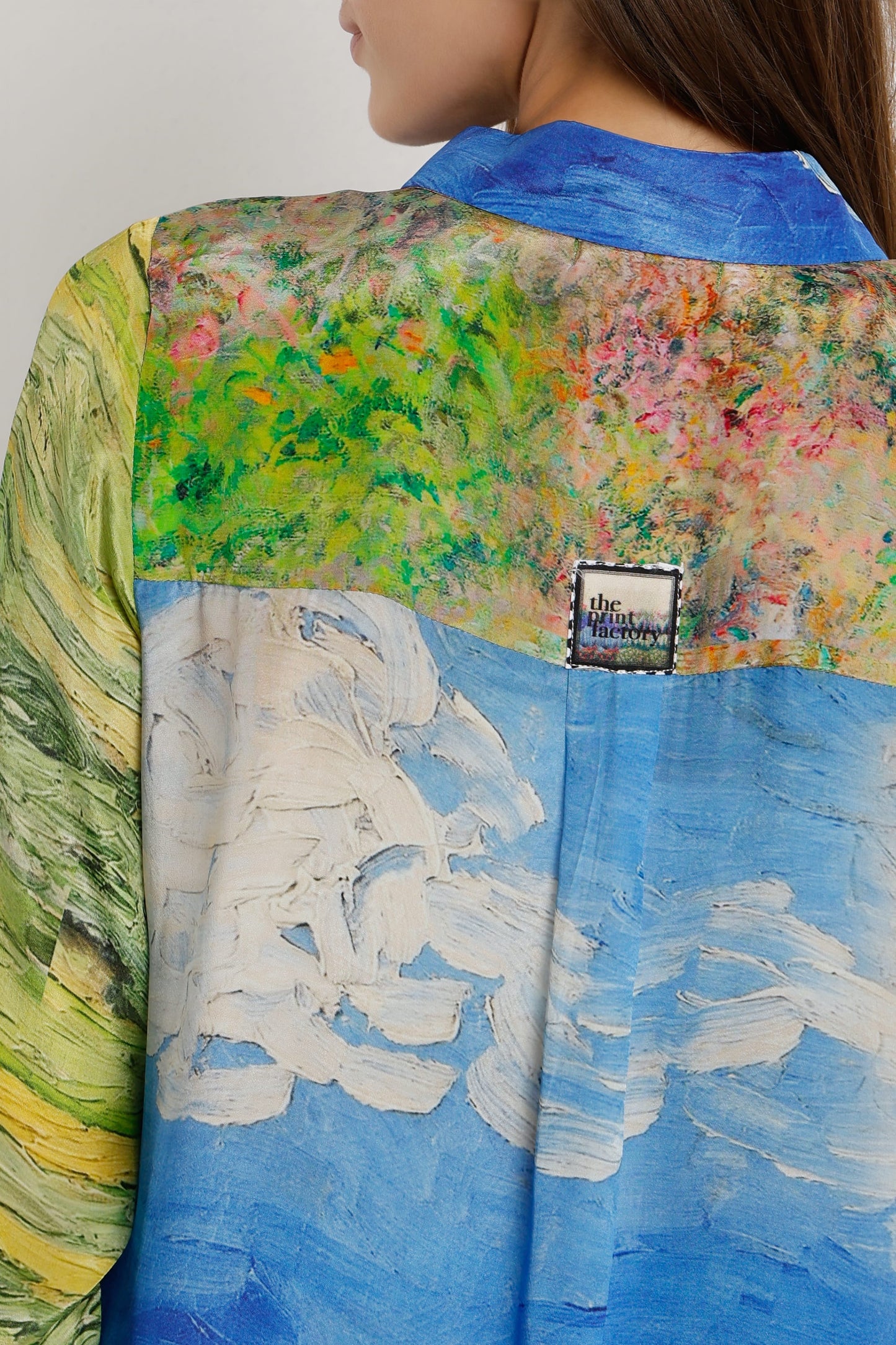 A close-up of a model wearing a silk shirt with an artistic, painterly print inspired by Van Gogh. The shirt features a blue sky and white cloud pattern on the lower half, a green and yellow landscape on the left, and a colorful floral field on the upper right.