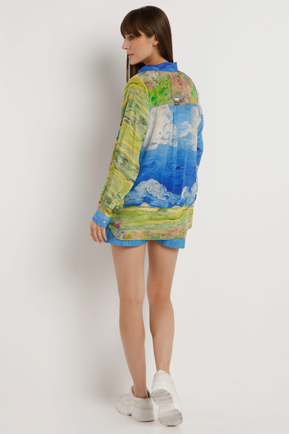 A woman modeling a long-sleeve button-up shirt with a colorful landscape print, shown from the back against a plain light gray studio background.