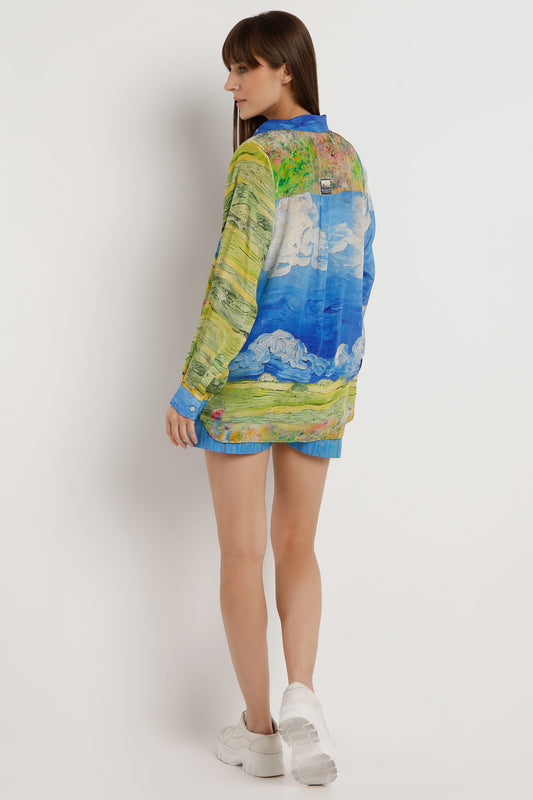 A woman modeling a long-sleeve button-up shirt with a colorful landscape print, shown from the back against a plain light gray studio background.