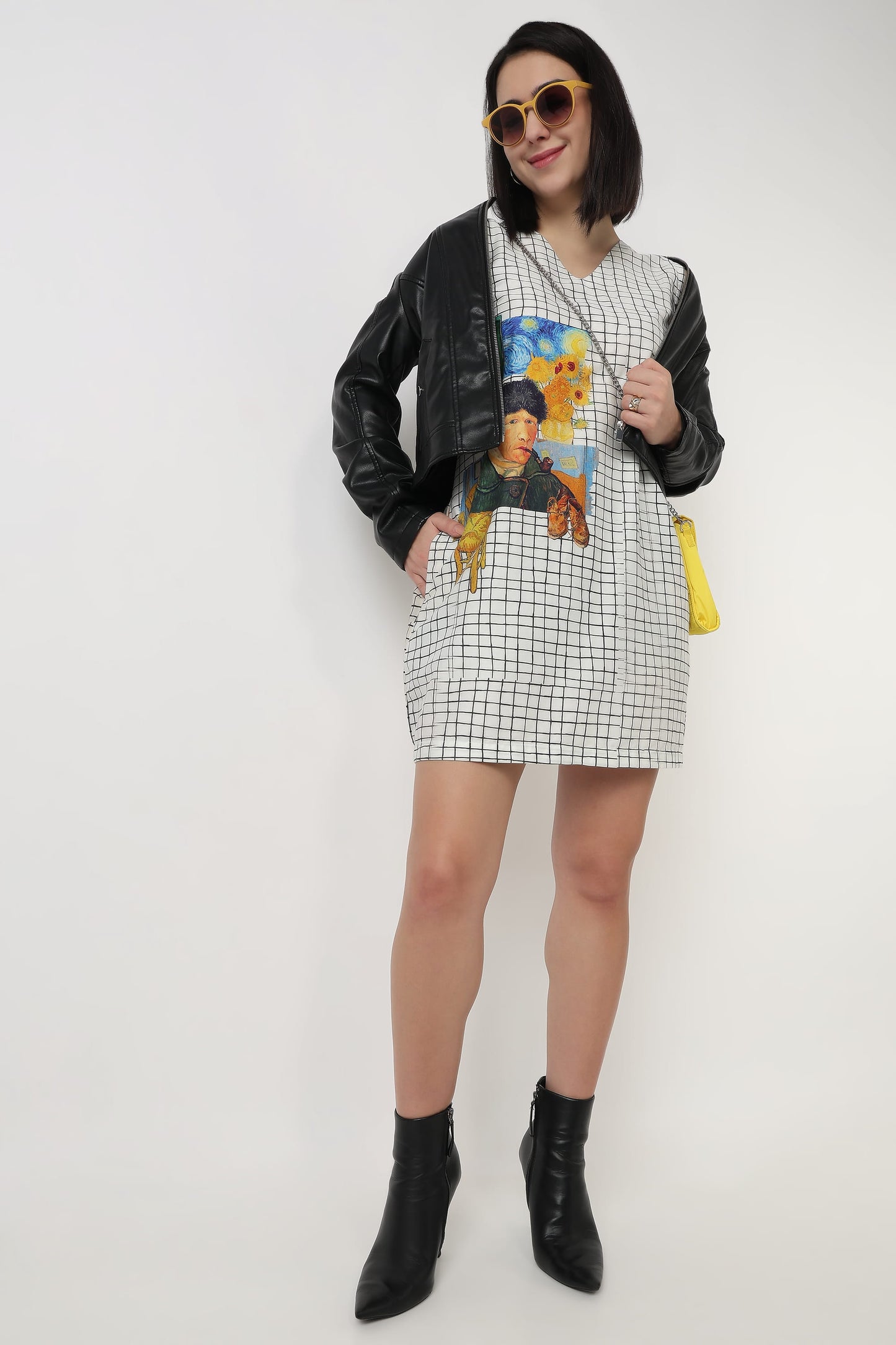 A woman modeling a black and white checkered mini dress with a colorful graphic print on the front, styled with a black faux-leather jacket, black ankle boots, and a small yellow handbag against a plain white background.