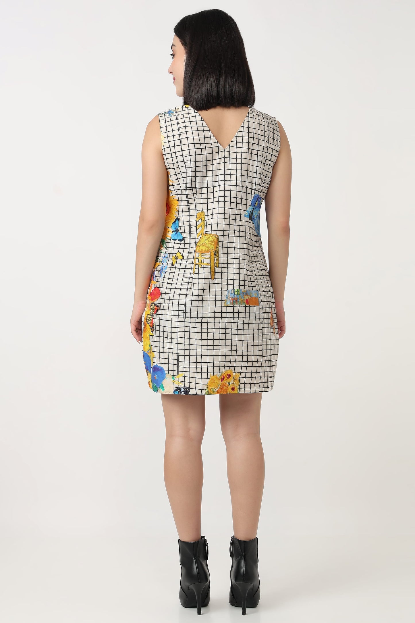 A woman viewed from the back wearing a sleeveless mini dress with a black and white grid pattern and colorful giraffe illustrations, paired with black ankle boots, against a plain light background.