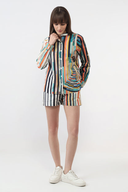 A woman modeling a matching two-piece striped lounge set consisting of a long-sleeve button-up shirt and shorts, featuring a multicolor vertical stripe pattern with a central graphic of a woman in a dress, paired with white sneakers on a light gray studio background.