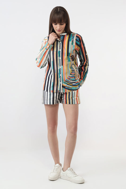 A woman modeling a matching two-piece striped lounge set consisting of a long-sleeve button-up shirt and shorts, featuring a multicolor vertical stripe pattern with a central graphic of a woman in a dress, paired with white sneakers on a light gray studio background.