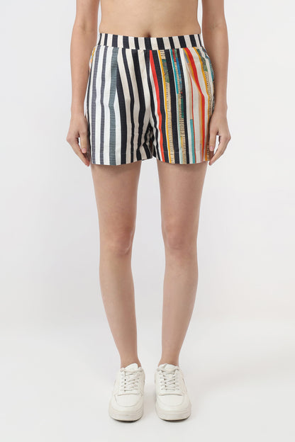 A model from the waist down wearing striped boxer-brief style shorts with vertical black, white, blue, red, orange, and yellow stripes, paired with white sneakers on a light gray studio background.