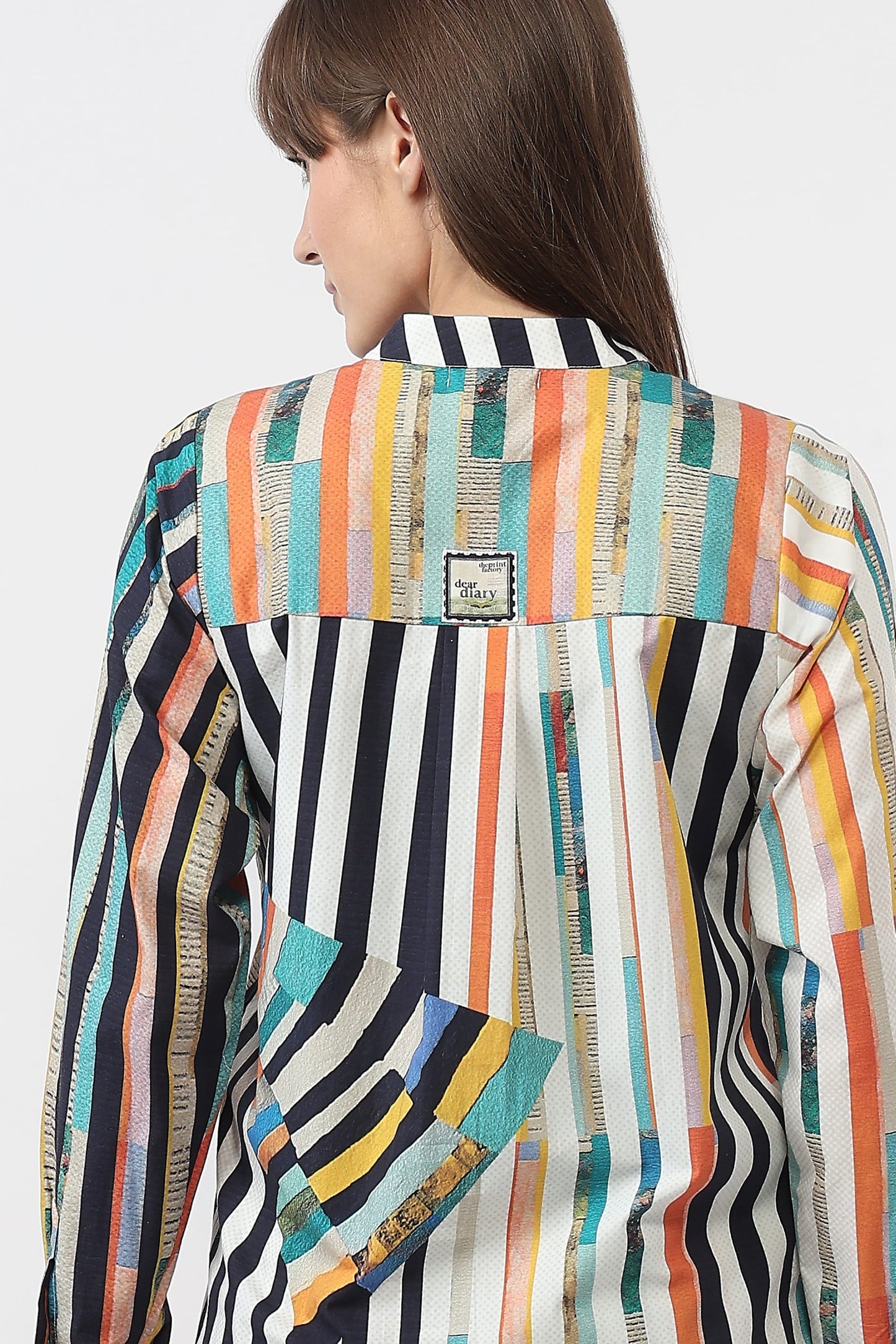 A woman shown from the back wearing a long-sleeve button-up shirt with a vibrant multicolor vertical stripe pattern in orange, teal, yellow, black, and white. A small square brand patch is visible on the upper back.
