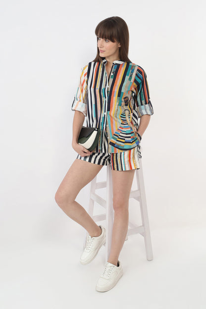 A woman models a matching two-piece set consisting of a short-sleeve button-up shirt and shorts, both featuring a vibrant multicolor vertical stripe pattern with a central graphic illustration. She is seated on a white stool against a plain white studio background, holding a black and white clutch bag.