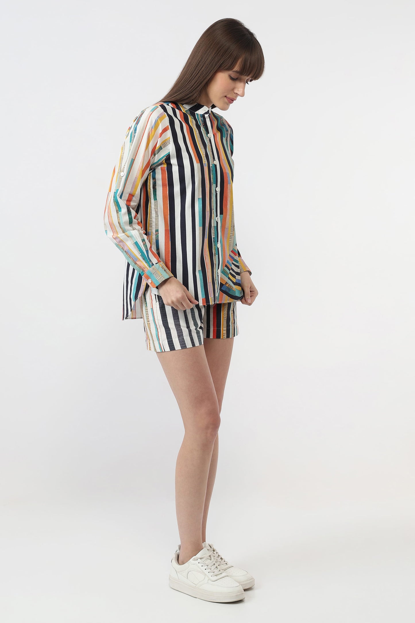 A woman modeling a matching two-piece set consisting of a long-sleeve button-up shirt and shorts, both featuring a vertical multicolor stripe pattern on a white base, paired with white sneakers against a plain white studio background.