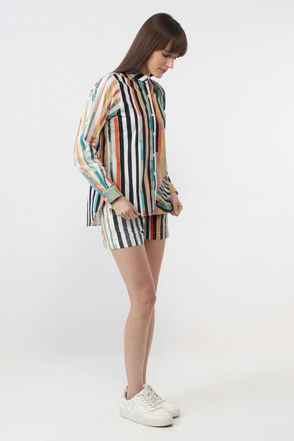 A woman modeling a matching two-piece set consisting of a long-sleeve button-up shirt and shorts, both featuring a vertical multicolor stripe pattern on a white base, paired with white sneakers against a plain white studio background.